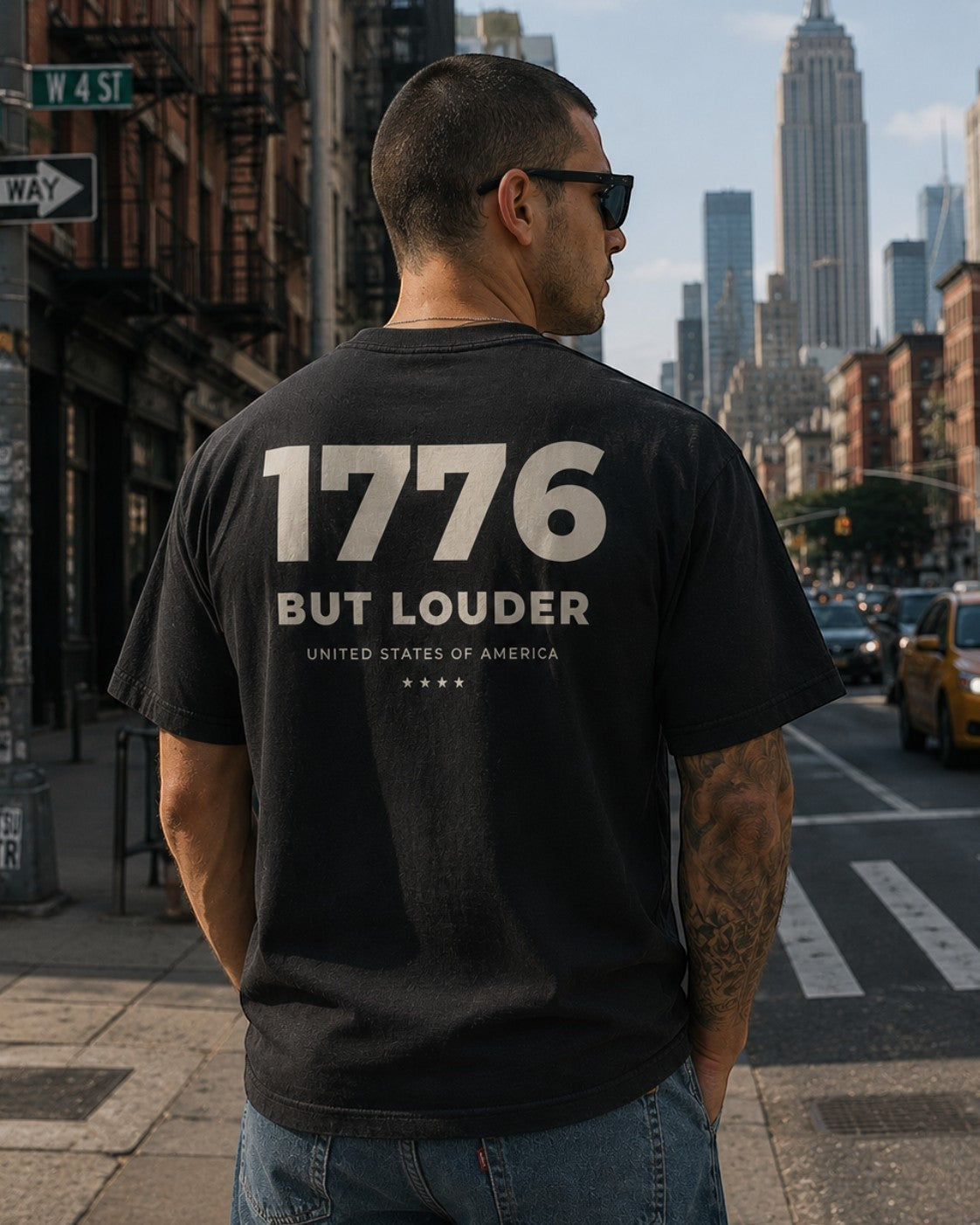 Americana black crewneck t-shirt with 1776 But Louder graphic on the back, worn by a man on a city street.