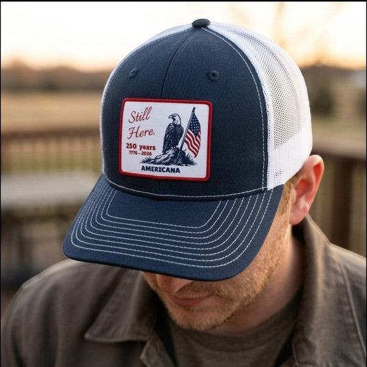 Americana Still Here trucker hat with navy blue front, white mesh back, and eagle American flag patch on a man.